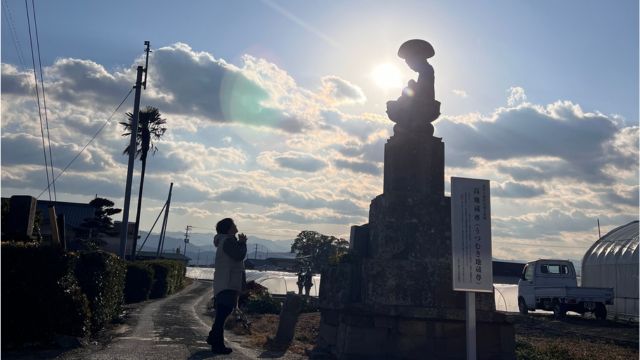 夕日を背に立つお地蔵さんに、手を合わせる人
